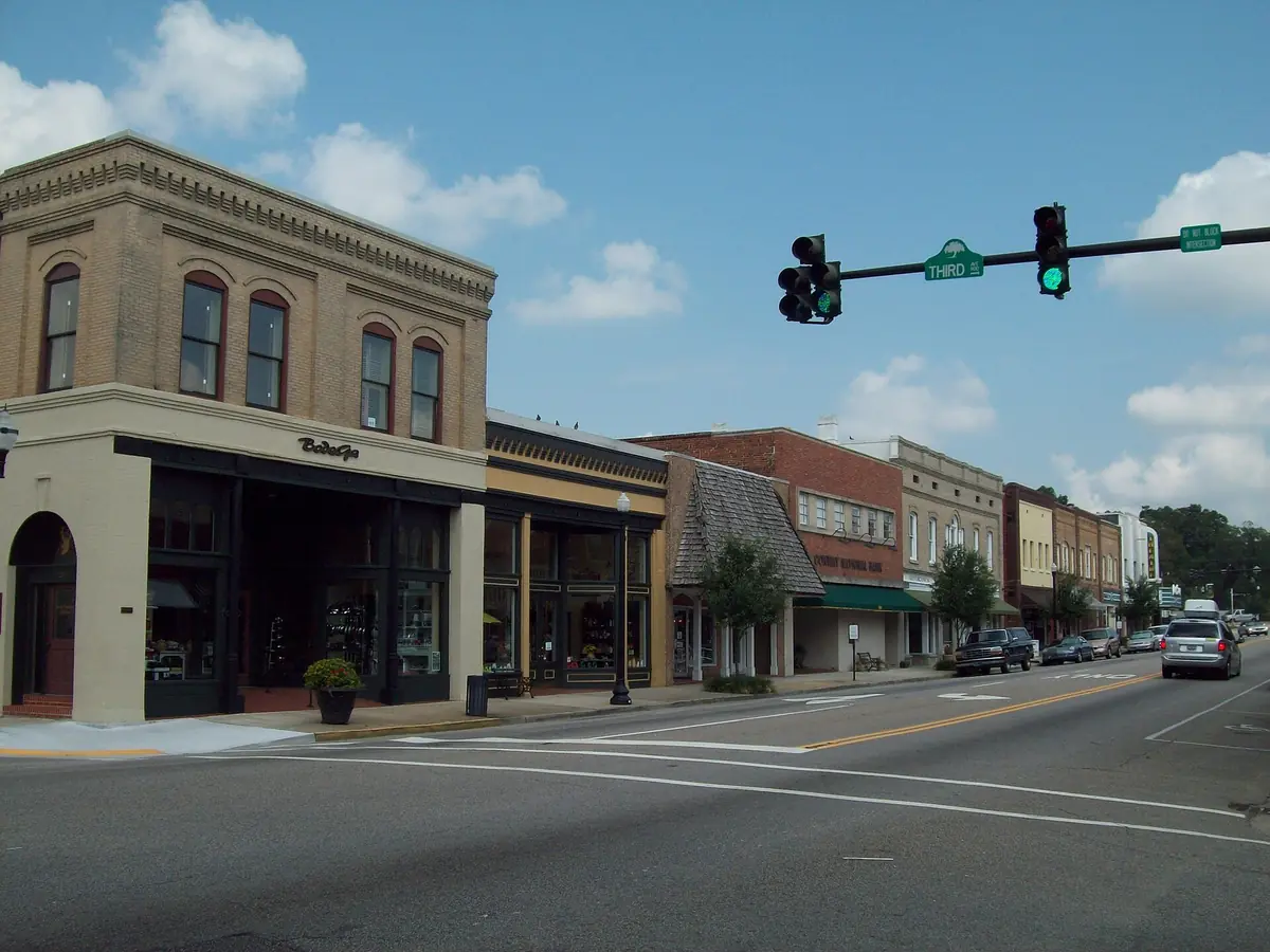 Main Street in downtown Conway, South Carolina, with a row of two-story brick and stone shop buildings, street trees and parked cars under a blue sky
