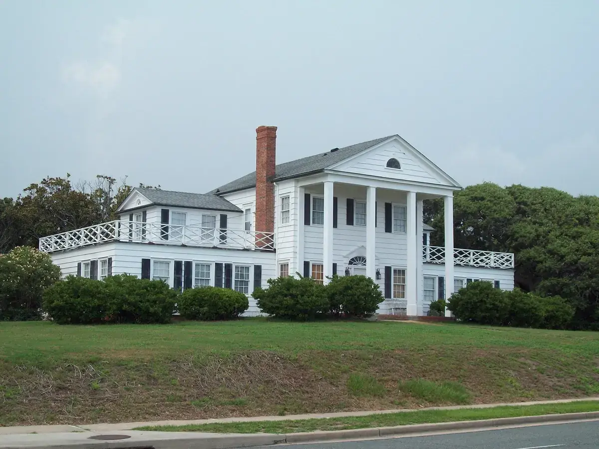 An older two-story white wood house with tall columns, black shutters and a brick chimney, on a grassy rise above North Ocean Boulevard in Myrtle Heights