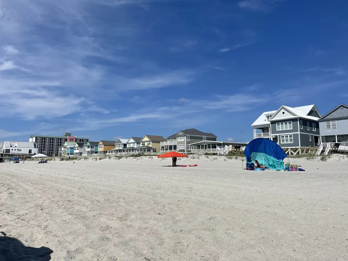A row of two- and three-story beach houses in blue, yellow and gray behind low dunes, seen across a wide sand beach under a blue sky at Surfside Beach